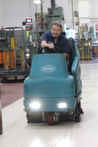 A crew member on a blue riding floor scrubber performing general cleaning.